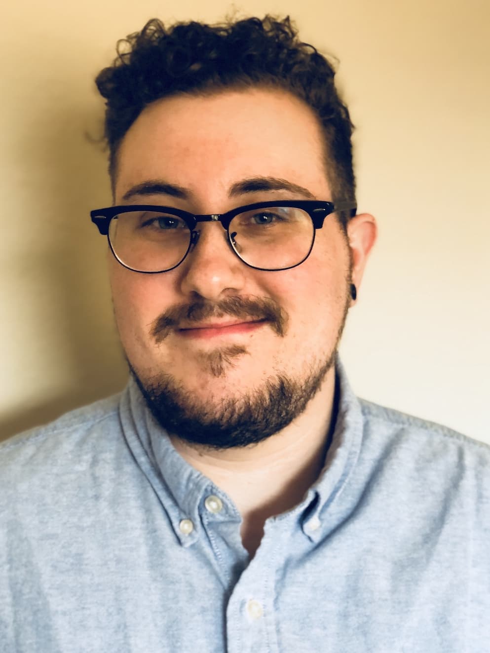 A picture of Andy, a white man, smiling at the camera. He has short, curly hair, dark-rimmed glasses, and gauges in his ears.